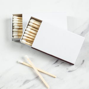 May include: White matchboxes with brown edges, each filled with wooden matches with white tips. Two matches are placed in front of the boxes. The matches and boxes are on a white marble surface.