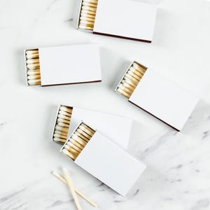 May include: Several white matchboxes with brown edges are scattered on a white marble surface. Each box is open, revealing rows of wooden matches with white tips. Two loose matches are also visible, suggesting a focus on home decor or fire-starting supplies.