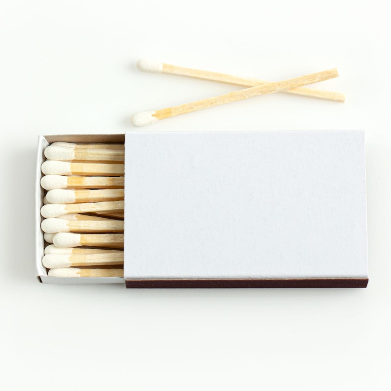 May include: A white matchbox with a brown striking surface, partially open to reveal a row of wooden matches with white tips. Two matches are placed on top, creating a simple, clean composition. The matches are made of wood.
