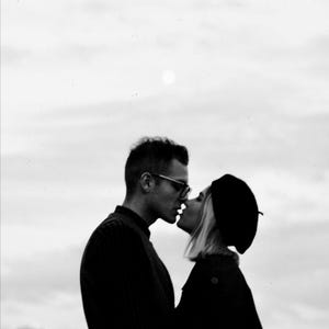 May include: Black and white photograph of two people kissing. The man wears glasses and a dark shirt. The woman wears a beret and a dark coat. The background is a cloudy sky.