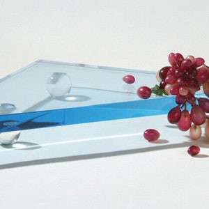 May include: A clear glass tray with a blue stripe and rounded edges. The tray is supported by three clear glass balls. A bunch of red grapes with green leaves is on the tray.