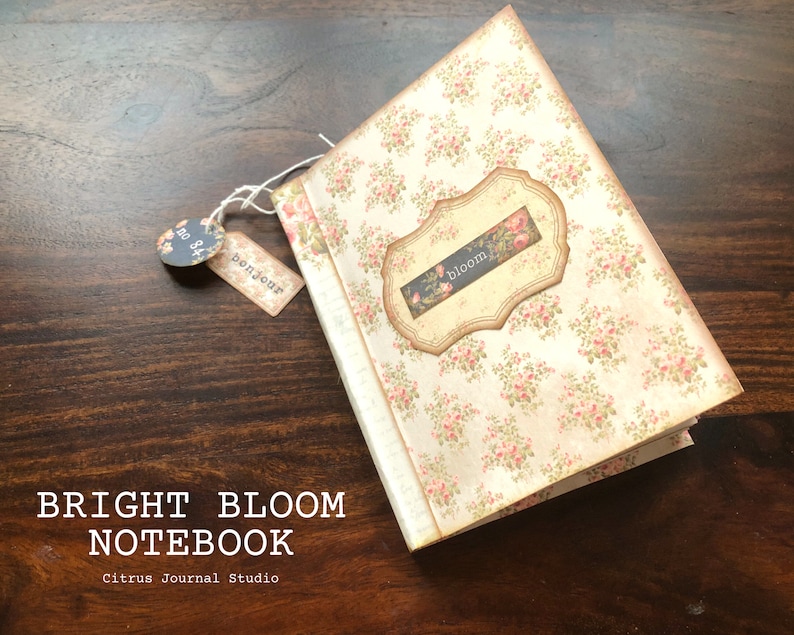 May include: A small, white notebook with a floral pattern and a label that says "bloom". The notebook has a string attached to it with a tag that says "bonjour".
