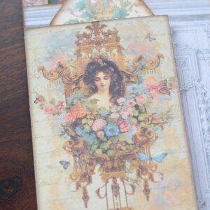 May include: A vintage-style illustration of a woman with long brown hair, surrounded by pink and blue flowers, in a gold ornate frame. The image is on a light beige background with a faded, vintage look.