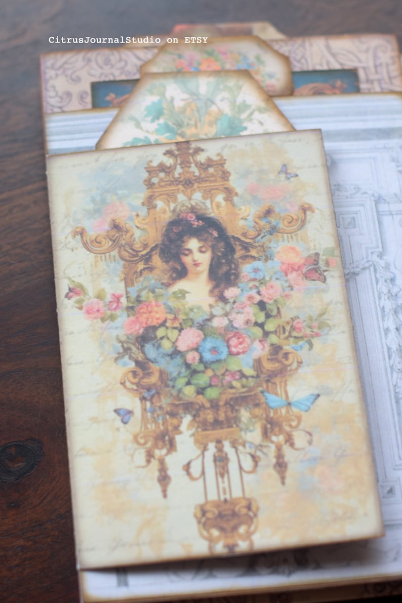 May include: A vintage-style illustration of a woman with long brown hair, surrounded by pink and blue flowers, in a gold ornate frame. The image is on a light beige background with a faded, vintage look.