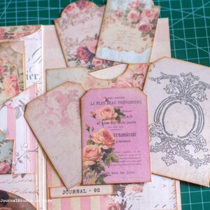 Pu&ograve; includere: Una collezione di efemeridi di carta in stile vintage, tra cui tag e inserti di diario. I pezzi presentano motivi floreali di rose in tonalit&agrave; rosa e crema, con alcuni tag che mostrano testo francese e decorazioni decorative. "JOURNAL 92" &egrave; stampato su una piccola etichetta.