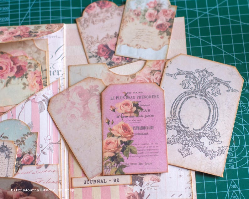 Pu&ograve; includere: Una collezione di efemeridi di carta in stile vintage, tra cui tag e inserti di diario. I pezzi presentano motivi floreali di rose in tonalit&agrave; rosa e crema, con alcuni tag che mostrano testo francese e decorazioni decorative. "JOURNAL 92" &egrave; stampato su una piccola etichetta.