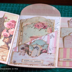 Pu&ograve; includere: Un diario fatto a mano in stile vintage con una combinazione di colori rosa e crema. Il diario presenta motivi floreali, tra cui rose, e i testi "Vintage Garden" e "Journal - 92". Il diario aperto rivela pi&ugrave; tasche e inserti decorativi.