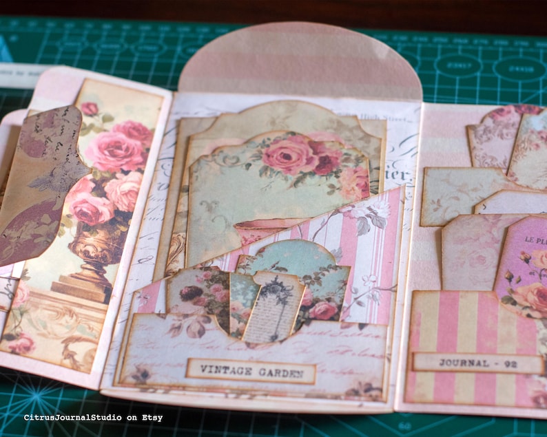 Pu&ograve; includere: Un diario fatto a mano in stile vintage con una combinazione di colori rosa e crema. Il diario presenta motivi floreali, tra cui rose, e i testi "Vintage Garden" e "Journal - 92". Il diario aperto rivela pi&ugrave; tasche e inserti decorativi.