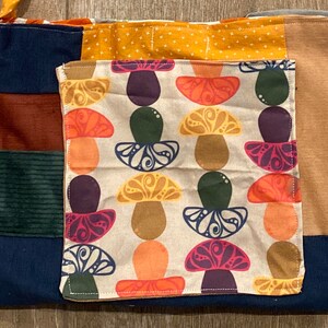 May include: A blue, brown, green, and yellow patchwork tote bag with a white fabric pocket featuring a mushroom pattern. The pocket has a brown stitched border.