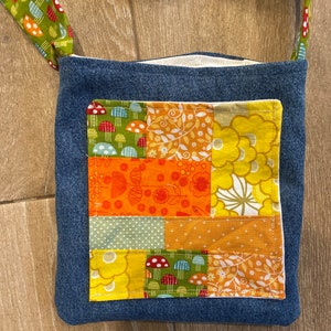 May include: A small denim bag with a patchwork design. The patchwork is made up of different fabrics with floral and mushroom patterns in shades of yellow, orange, green, and red.
