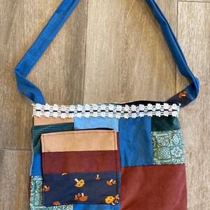 May include: A patchwork tote bag with a blue strap and white lace trim. The bag is made of different fabrics in shades of blue, green, brown, and red. It has a large pocket on the front.