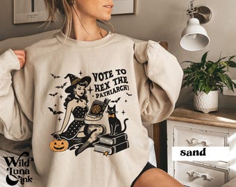 May include: A beige sweatshirt with a graphic of a witch reading a book with a black cat sitting next to her. The text on the sweatshirt reads "Vote to Hex the Patriarchy".