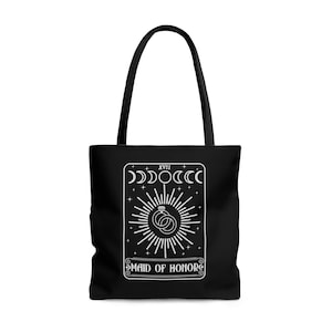 May include: Black tote bag with a white design of a tarot card. The card is labeled "XVII" and "Maid of Honor". The card features a stylized sun with rings and a diamond.