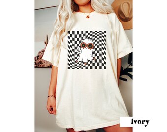 May include: Ivory-coloured t-shirt with a cartoon ghost wearing sunglasses and giving a peace sign, set against a black and white chequered background. The shirt has a relaxed fit and short sleeves. The word "ivory" is printed at the bottom.