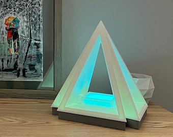 May include: A white pyramid-shaped lamp with a light blue glow emanating from the base. The lamp has a rectangular base and a clear glass insert in the centre.