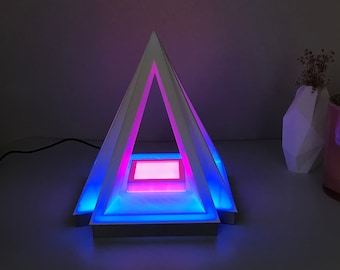 May include: A white pyramid-shaped lamp with a blue and pink light emanating from the base. The lamp is on a white surface.