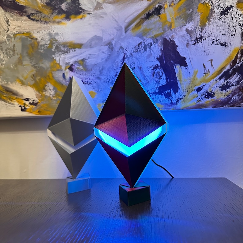 May include: Two Ethereum cryptocurrency logo lamps, one is black with a blue light and the other is silver. The lamps are on a dark wood surface.