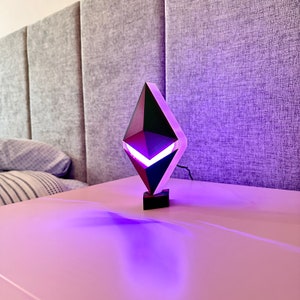 May include: A black and purple geometric lamp shaped like a diamond with a white light inside. The lamp is sitting on a white table.
