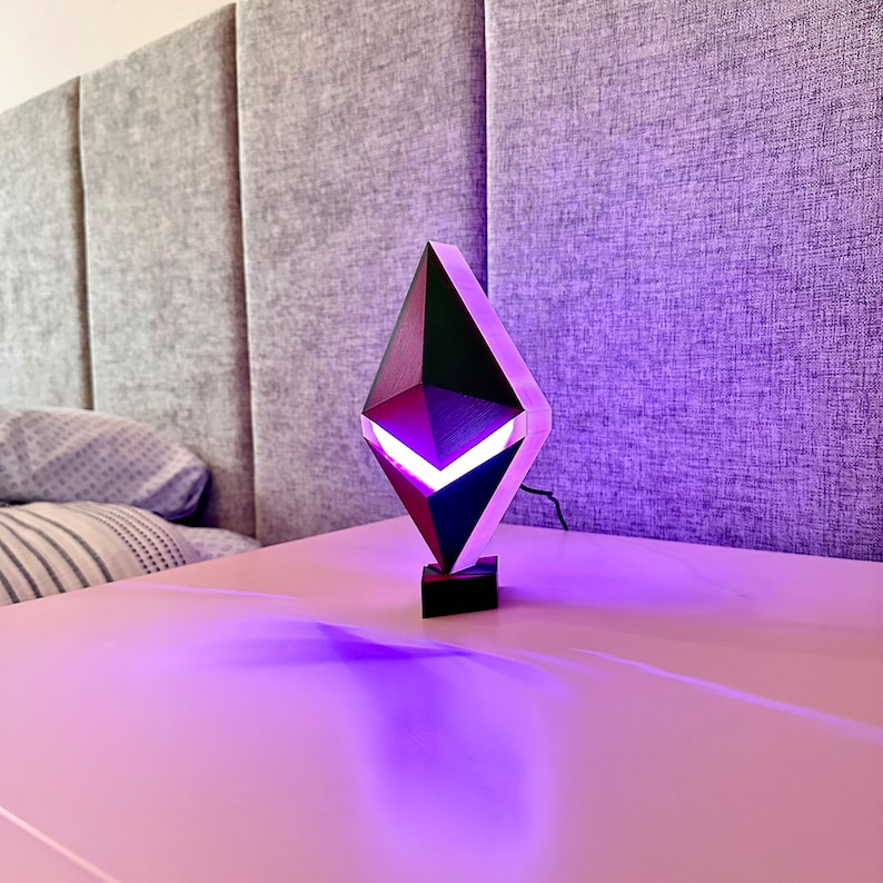 May include: A black and purple geometric lamp shaped like a diamond with a white light inside. The lamp is sitting on a white table.