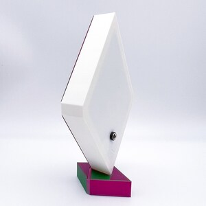 May include: A white diamond-shaped lamp with a green and magenta base. The lamp has a cord and plug on the bottom.
