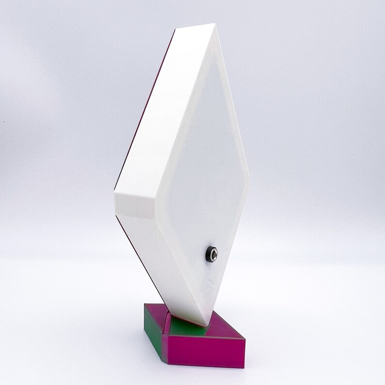 May include: A white diamond-shaped lamp with a green and magenta base. The lamp has a cord and plug on the bottom.