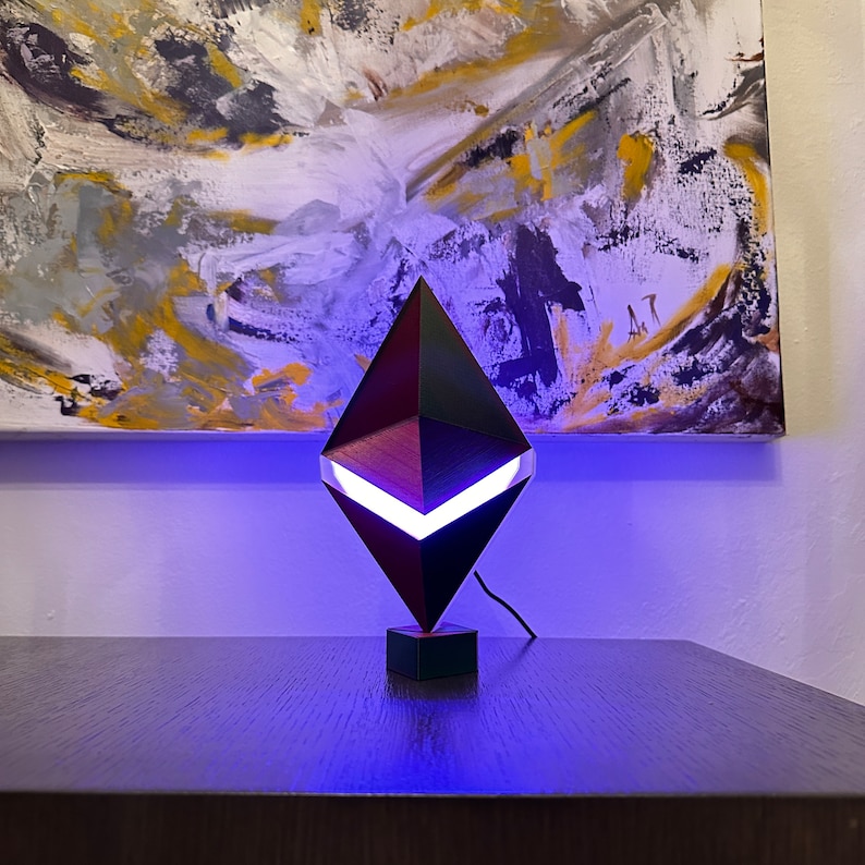 May include: A black Ethereum cryptocurrency lamp with a white light shining from the centre. The lamp is on a dark wooden surface.