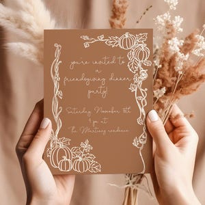 May include: A brown invitation card with white text and pumpkin illustrations. The card reads "you're invited to a friendsgiving dinner party!" with the date and time. The card is held in front of a neutral background.