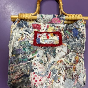 May include: A handmade patchwork tote bag with a bamboo handle. The bag is made from various fabric scraps in shades of blue, white, pink, and red. A red label with white text is sewn on the front. The bag is on a purple surface.