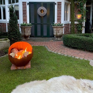 Könnte beinhalten: Herbstliche Gartenszene mit einem brennenden Kürbis-Feuerkorb auf einem Holzsockel, auf Rasen. Im Hintergrund ein Backsteingebäude mit grünen Fensterläden und einem Kranz. Ein Backsteinweg und Grün umgeben die Szene.