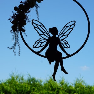 Può includere: Una silhouette di fata in metallo nero appesa a un cerchio con una ghirlanda di fiori. La fata è seduta sul cerchio con le ali spiegate.