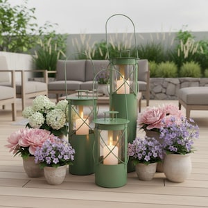 Può includere: Tre lanterne in metallo verde salvia con inserti cilindrici in vetro e manici ad arco, ciascuna contenente una candela a colonna accesa. Le lanterne sono disposte tra vari fiori in vaso, tra cui ortensie bianche e peonie rosa.