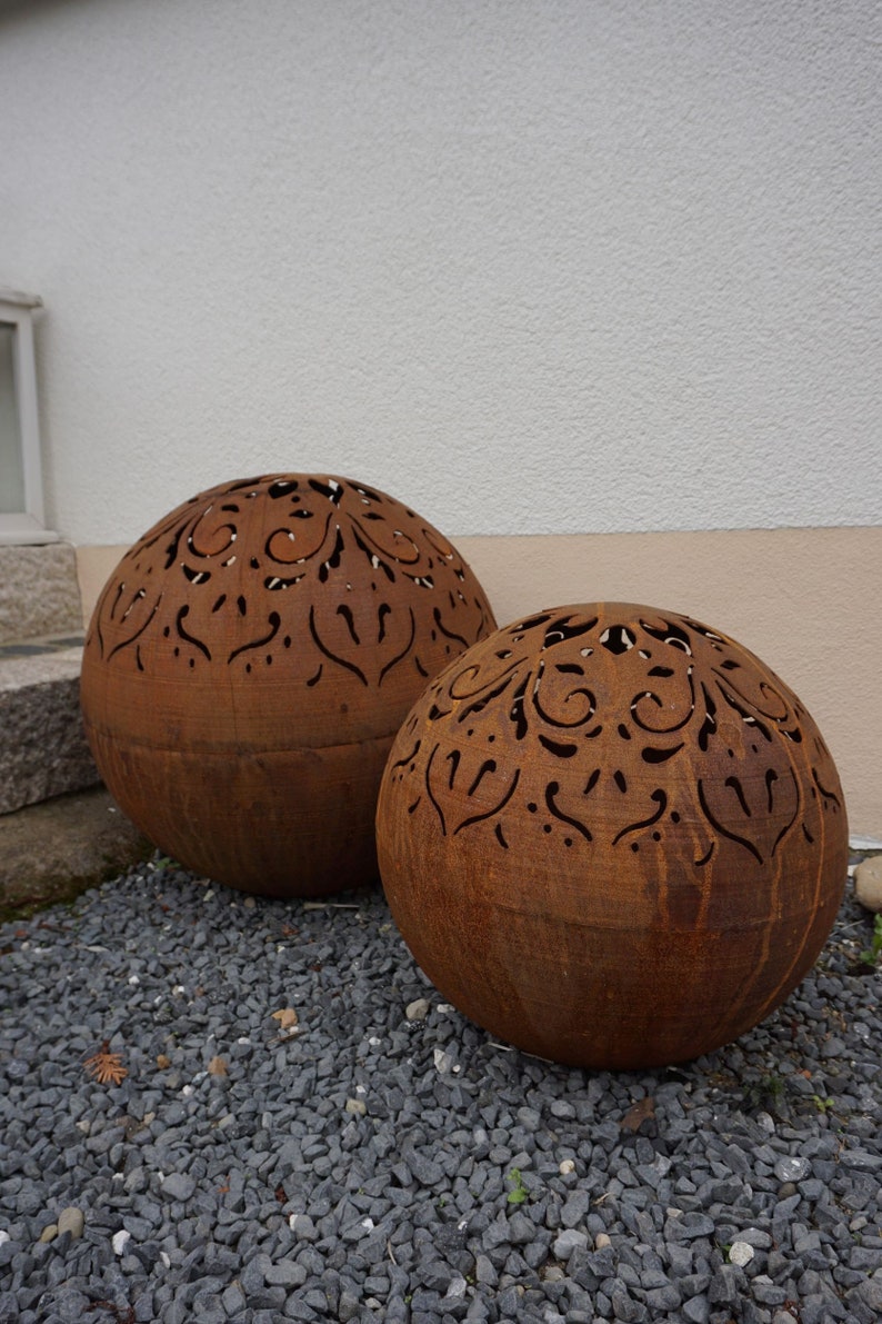 Puede incluir: Dos grandes esferas de metal oxidado con intrincados dise&ntilde;os recortados. Las esferas est&aacute;n sentadas sobre un lecho de peque&ntilde;as gravas grises.