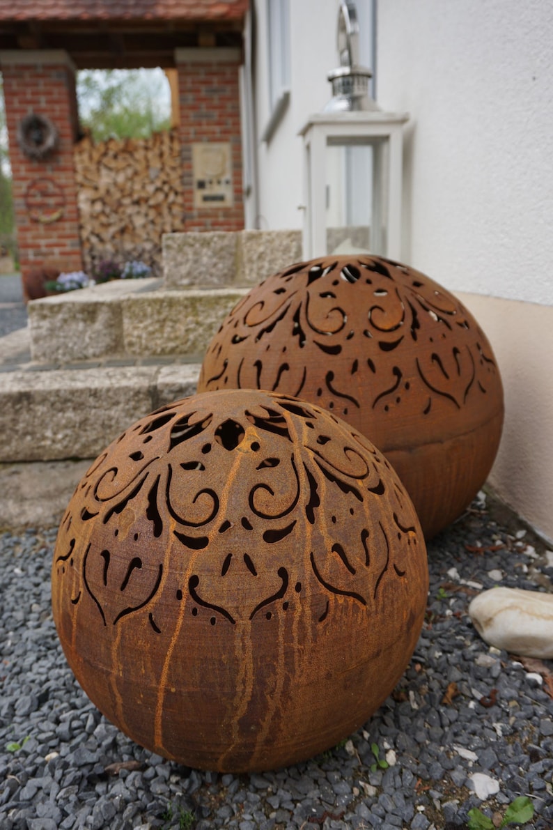 Puede incluir: Dos grandes esferas de metal oxidado con intrincados dise&ntilde;os recortados. Las esferas est&aacute;n sentadas sobre un lecho de peque&ntilde;as piedras grises.