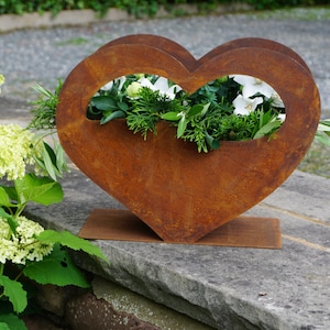 Könnte beinhalten: Ein rostiger, herzförmiger Blumentopf aus Metall mit einem Stand, gefüllt mit grünen Pflanzen und weißen Blumen.