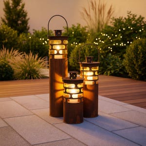 Peut inclure: Trois lanternes en métal rouillé de différentes hauteurs, dégageant une lueur chaleureuse. Chaque lanterne a un corps cylindrique avec des découpes rectangulaires et une poignée. Elles sont posées sur un patio en pierre, avec de la verdure en arrière-plan.