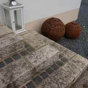 Puede incluir: Un conjunto de dos esferas met&aacute;licas decorativas con intrincados dise&ntilde;os recortados. Las esferas son de color marr&oacute;n oxidado y se colocan en un conjunto de escalones de piedra que conducen a un edificio blanco.