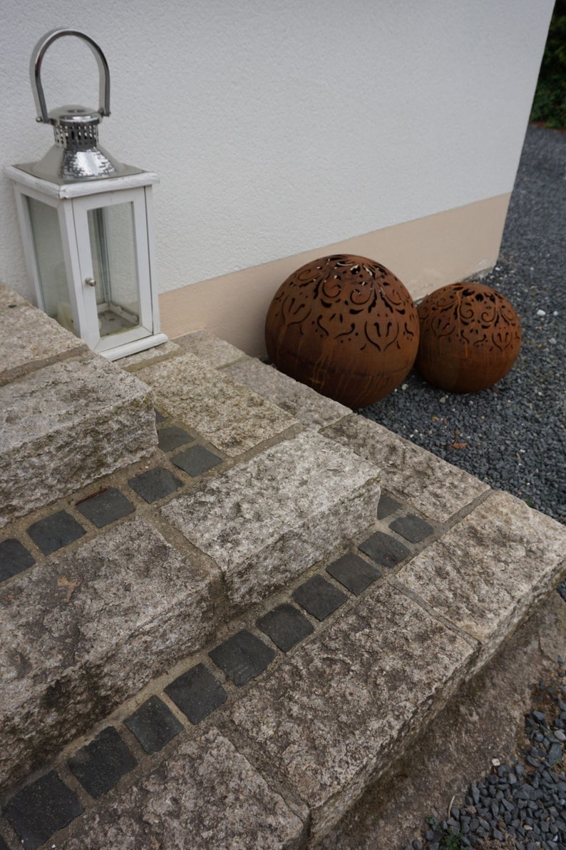 Puede incluir: Un conjunto de dos esferas met&aacute;licas decorativas con intrincados dise&ntilde;os recortados. Las esferas son de color marr&oacute;n oxidado y se colocan en un conjunto de escalones de piedra que conducen a un edificio blanco.