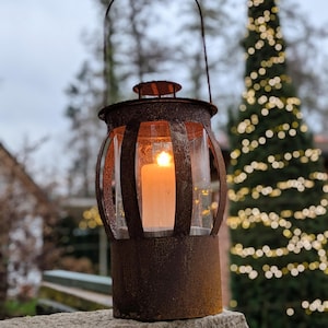 Peut inclure: Une lanterne cylindrique en métal vieilli avec une poignée. Elle est ornée de panneaux de verre transparents et d'une bougie blanche allumée à l'intérieur. En arrière-plan, un sapin de Noël flou avec des lumières et une surface en pierre.