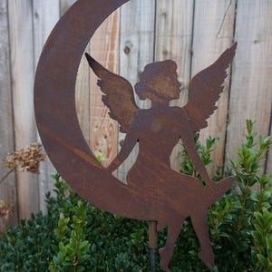 May include: A rustic metal garden stake featuring a silhouette of an angel seated on a crescent moon. The angel has outstretched wings and is set against a wooden fence and green foliage.