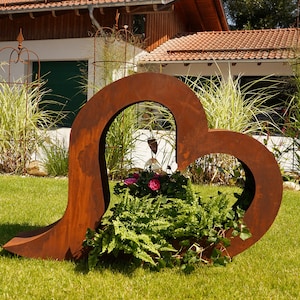 May include: A rusty metal heart-shaped planter with a fern and pink flowers growing inside. The planter is in a grassy area in front of a building.
