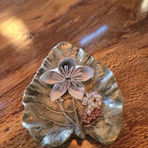 以下が含まれることがあります： ブロンズ仕上げの葉の形をした装飾的な皿には、紙の花、金の指輪、小さなピンクと白のビーズのクラスターが置かれています。 皿は木製の表面に置かれ、素朴な美しさを生み出しています。