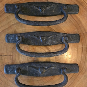 Könnte beinhalten: Drei rustikale, schwarze Metall-Schubladengriffe mit gehammerter Textur. Jeder Griff hat einen gebogenen Griff und eine rechteckige Rückplatte, die mit einem Longhorn-Stier-Emblem verziert ist. Geeignet für Möbel und Schränke.