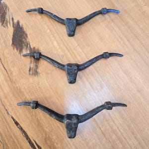 Könnte beinhalten: Drei rustikale, dunkle Metall-Stierhorn-Skulpturen, jede mit einem zentralen Kopf und gebogenen Hörnern. Die Hörner sind auf einer hellbraunen Holzoberfläche montiert, was einen auffälligen Kontrast erzeugt. Die Skulpturen sind wahrscheinlich Dekorationsartikel.