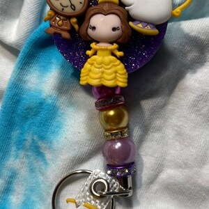 Pode incluir: Um porta-crachá com personagens de A Bela e a Fera. O porta-crachá tem um fundo roxo brilhante com Bela, Horloge e Sra. Potts. O porta-crachá tem um design de contas com uma alça branca e um pingente Chip.
