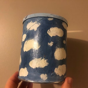 May include: A blue cylindrical container with a white painted design of clouds. The container has a light blue rim.