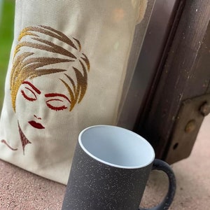 May include: A black and white mug with a speckled finish. The mug has a white interior and a black handle. The mug is sitting next to a white canvas tote bag with an embroidered image of a woman's face.