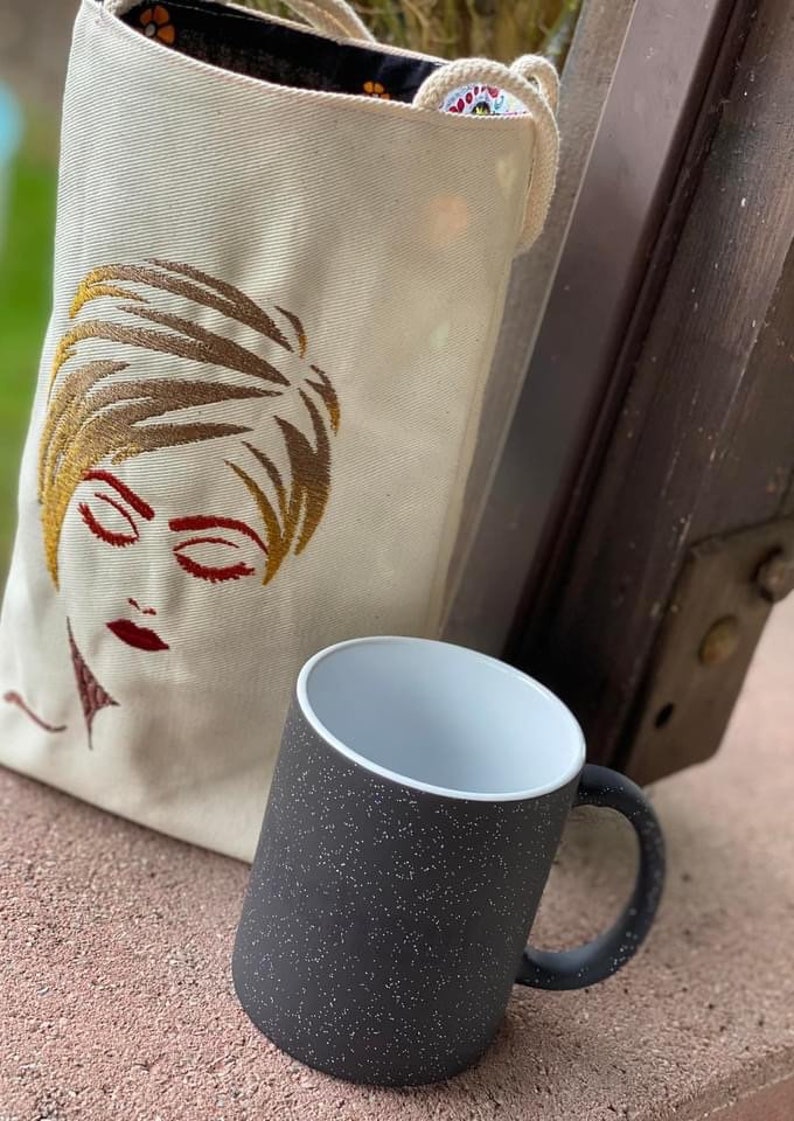 May include: A black and white mug with a speckled finish. The mug has a white interior and a black handle. The mug is sitting next to a white canvas tote bag with an embroidered image of a woman's face.