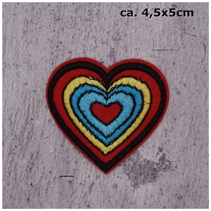 Könnte beinhalten: Ein roter herzförmiger Patch mit konzentrischen Kreisen aus gelber, blauer und roter Stickerei. Der Patch ist etwa 4,5 x 5 Zentimeter groß.