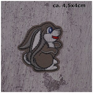 Könnte beinhalten: Ein grauer gestickter Aufnäher mit einem Cartoon-Kaninchen mit blauen Augen und rotem Mund. Der Aufnäher ist etwa 4,5 x 4 Zentimeter groß.