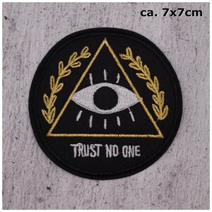 Könnte beinhalten: Schwarzer und goldener gestickter Aufnäher mit einem allsehenden Auge in einem Dreieck, umgeben von einem Kranz. Der Text "TRUST NO ONE" befindet sich unter dem Dreieck.